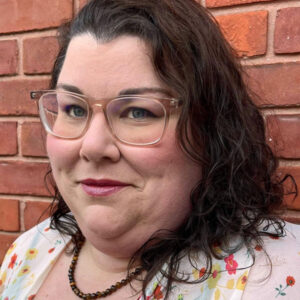 Woman with dark curly hair and glasses in front of a brick wall.